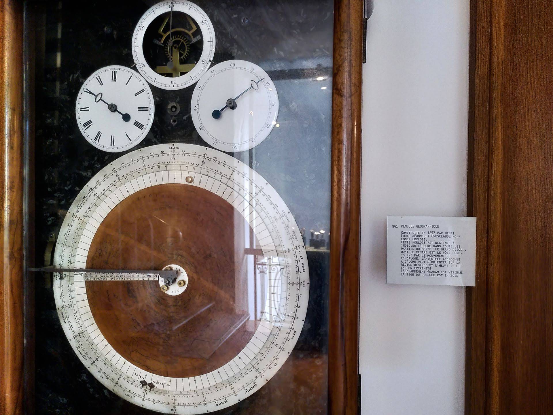 A complex time-keeping device. The text on the side reads "Pendule Geographique. Construite en 1857 par Henri Louis Jeanneret-Grosclaude horloger loclois. Cette horloge est destinée à indiquer l'heure dans toute les parties du monde."
