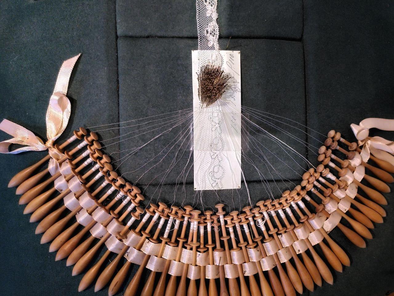 A lace ribbon interrupted in its creation, with a pin-board and array of Bobbins, on display